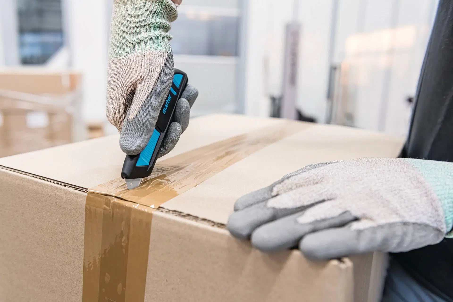 Hand mit Schutzhandschuh schneidet das Paketklebeband eines Kartons präzise mit dem MARTOR SECUNORM 300 Sicherheitsmesser von SICHERHEITSCUTTER.DE auf.