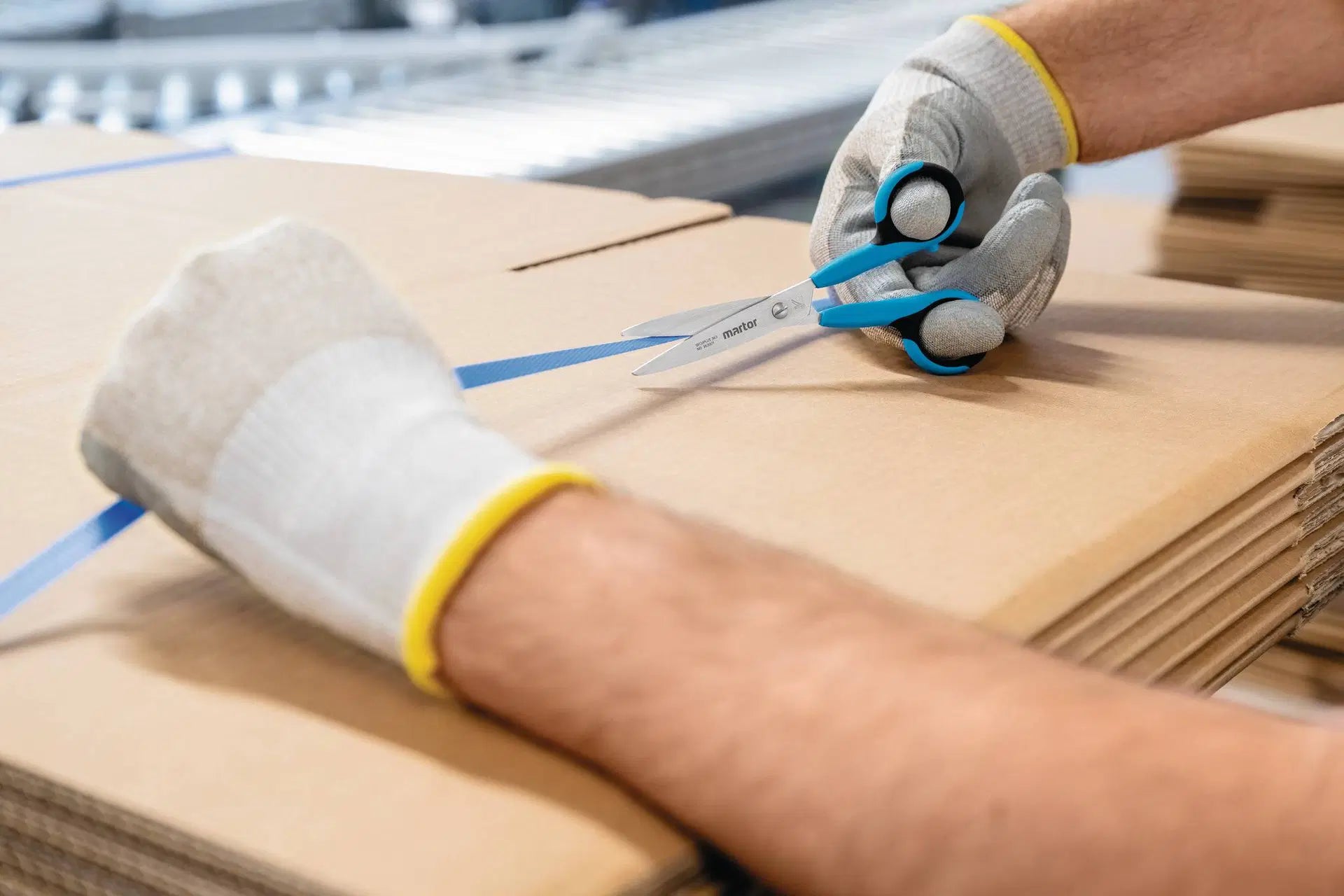 Hand mit Schutzhandschuh durchtrennt ein blaues Kunststoff-Umreifungsband präzise mit der MARTOR SECUPLUS 363 Sicherheits-Schere von SICHERHEITSCUTTER.DE.