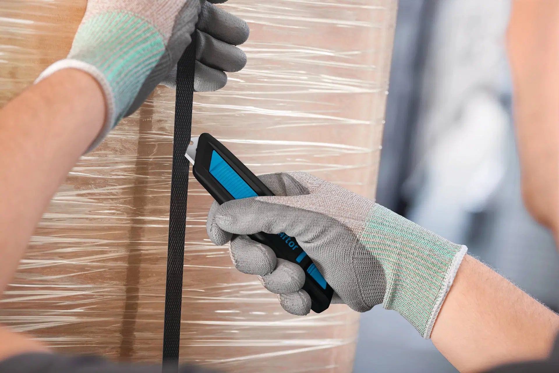 Hand mit Schutzhandschuh durchtrennt ein Kunststoff-Umreifungsband sicher mit dem MARTOR SECUNORM 300 Sicherheitsmesser von SICHERHEITSCUTTER.DE.