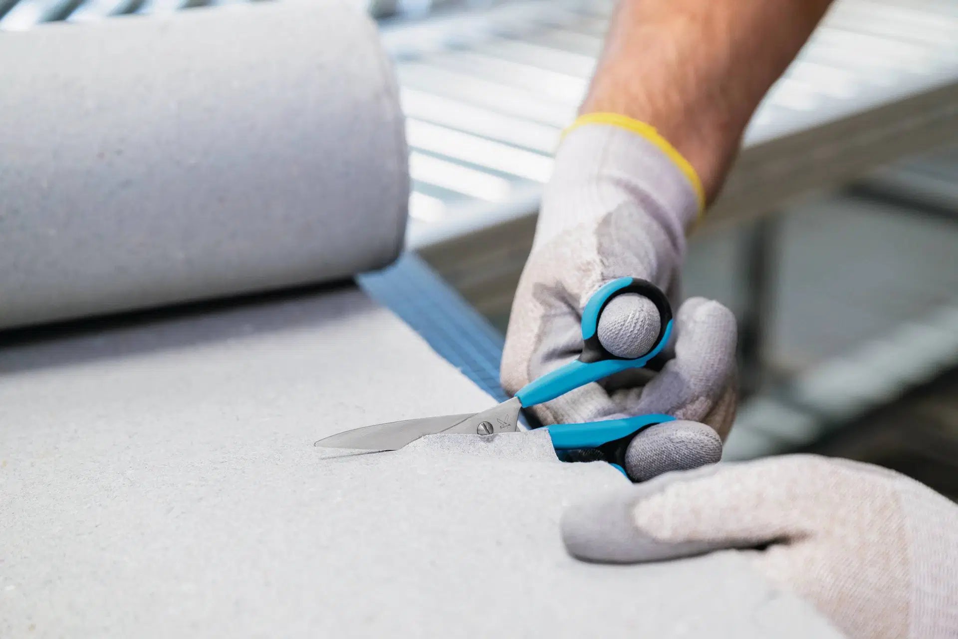 Hand mit Schutzhandschuh schneidet dickes Vlies oder Filzmaterial von einer Rolle präzise mit der MARTOR SECUPLUS 363 Sicherheits-Schere von SICHERHEITSCUTTER.DE.