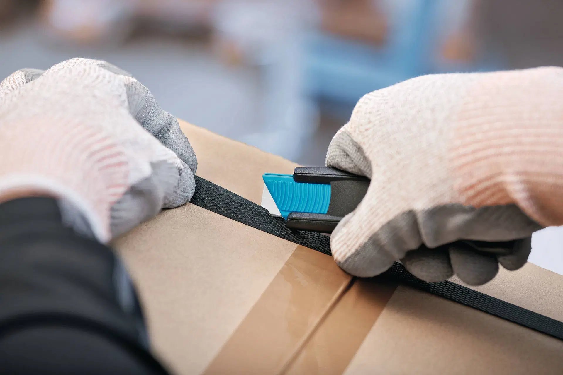 Hand mit Schutzhandschuh schneidet Wickelfolie auf einem Karton präzise mit dem handlichen MARTOR SECUNORM 175 Sicherheitsmesser von SICHERHEITSCUTTER.DE auf.